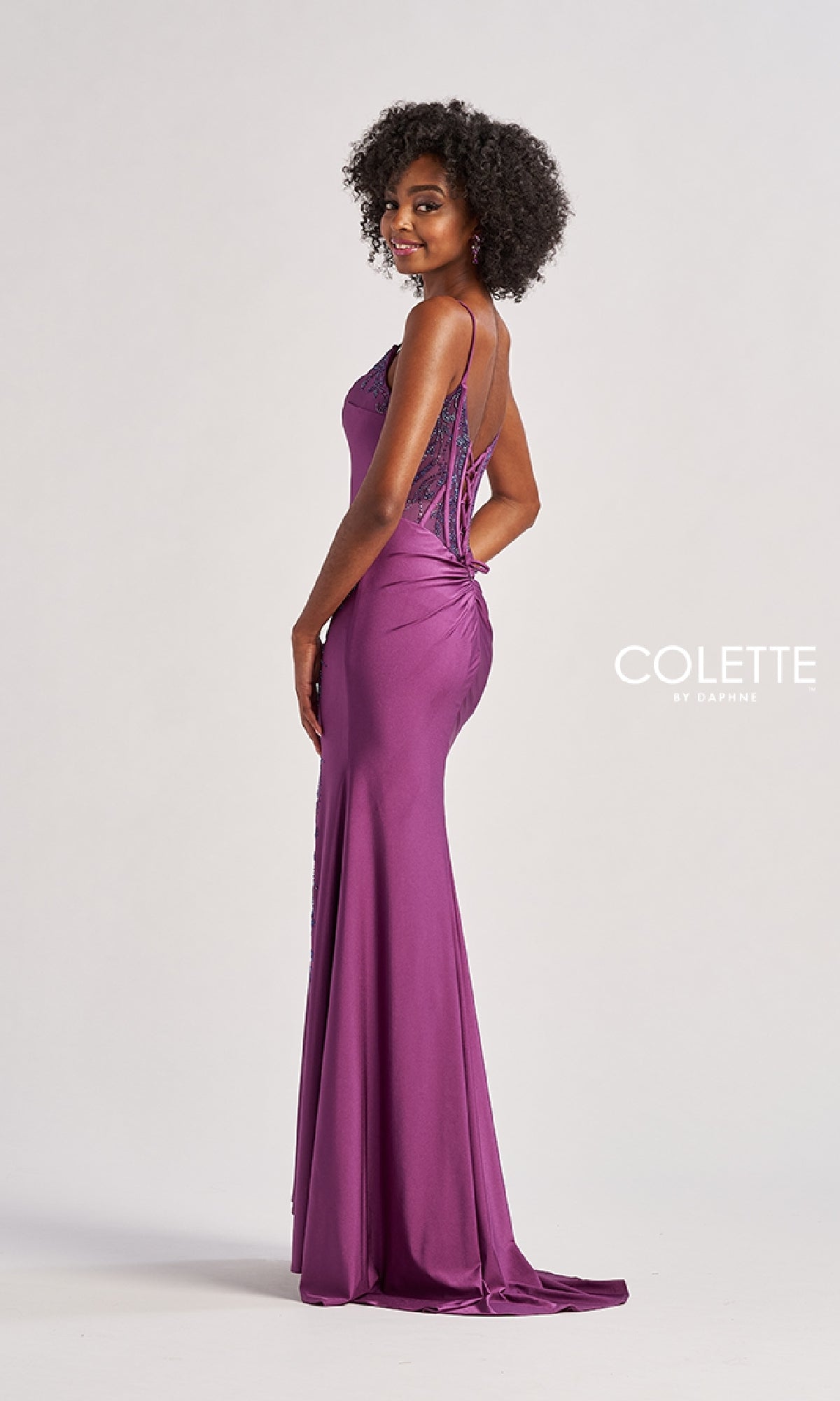 A woman stands sideways in the floor-length Colette CL8485 prom dress, a fitted purple gown with lace details, corset bodice, and lace-up back.